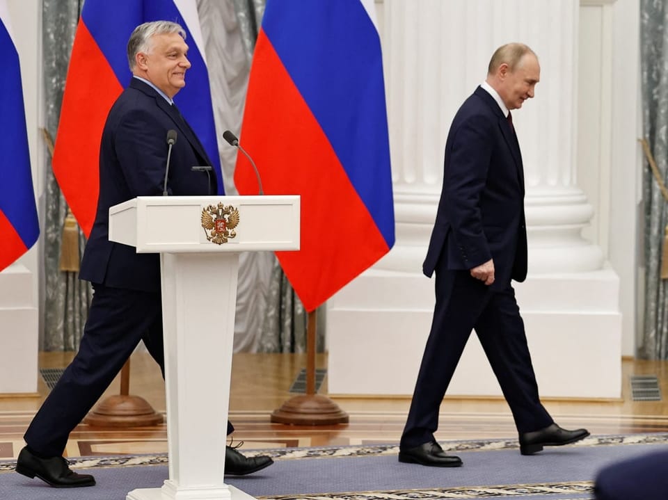 Zwei Männer in Anzügen bei einer offiziellen Veranstaltung mit russischen Flaggen im Hintergrund.