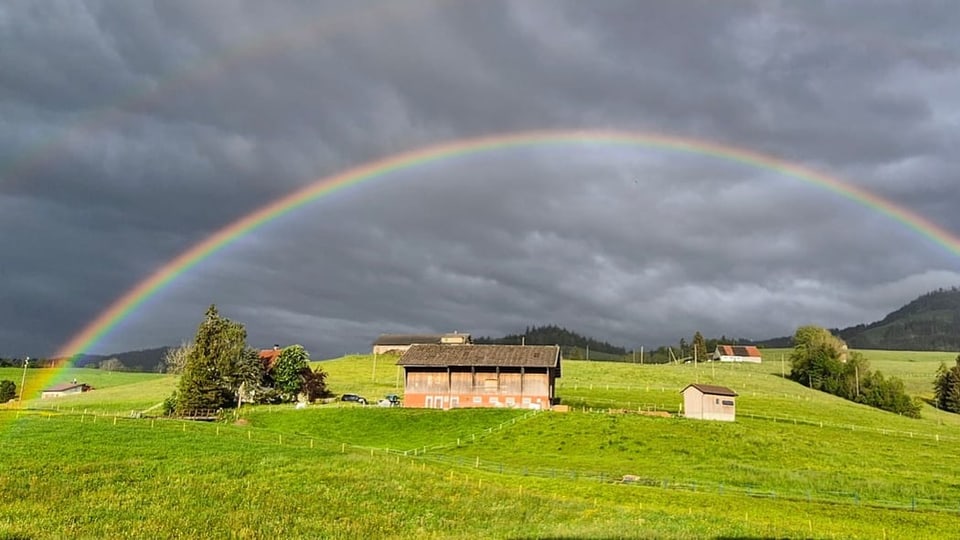 Regenbogen über grünem Feld und Gebäuden.
