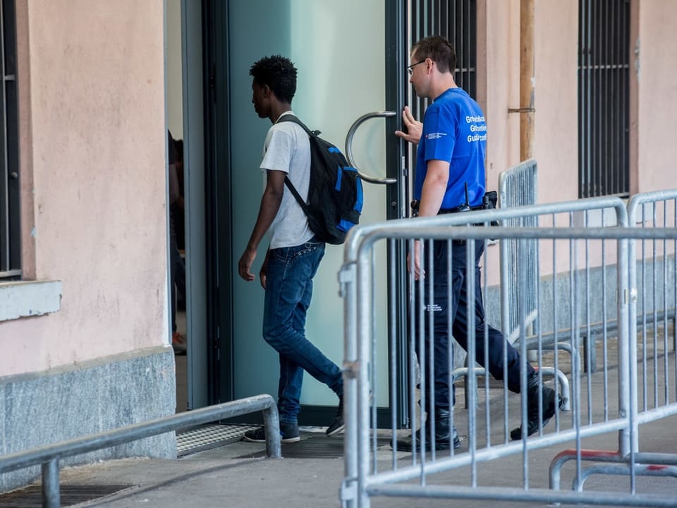 Mann mit Rucksack tritt in Gebäude ein, begleitet von Wachmann.