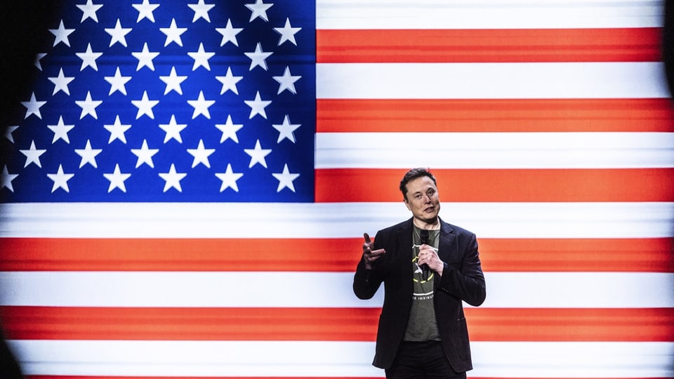 Mann mit T-Shirt und Anzug steht vor einer US-Flagge.