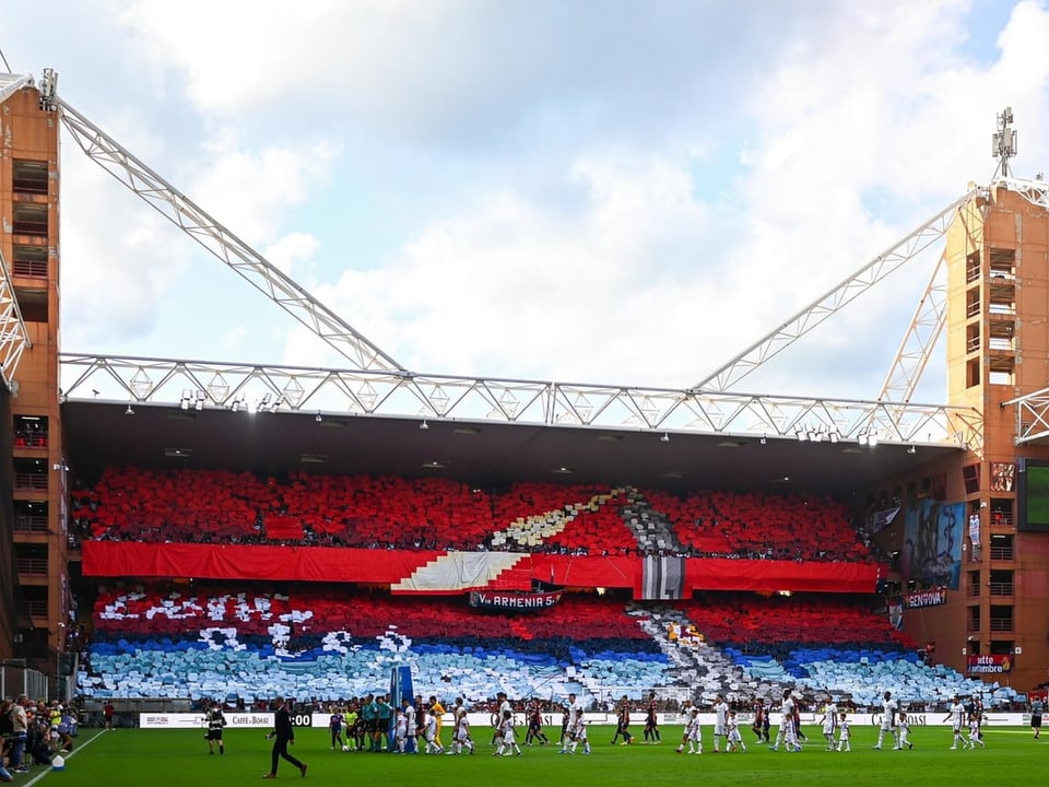 Fussballstadion mit gefüllten Tribünen, rot-blaues Banner und Mannschaft auf dem Spielfeld.