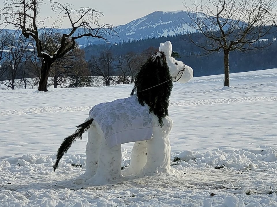 Verschneiter Park mit Pferde-Schneeskulptur und Bergen im Hintergrund.