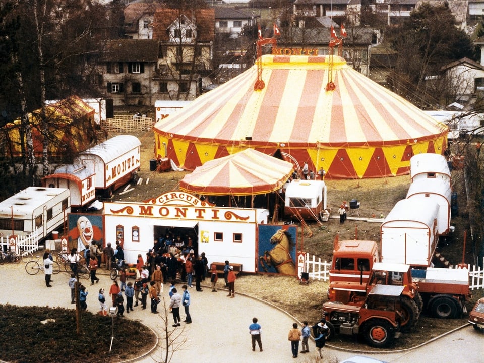 Luftaufnahme eines Zirkusplatzes mit rot-gelbem Zirkuszelt und Menschenmenge.