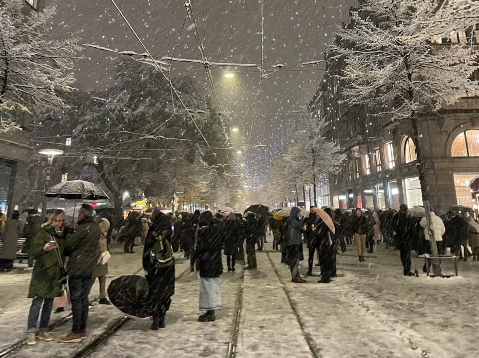 Menschenmenge bei Schneefall in einer beleuchteten Stadtstrasse.