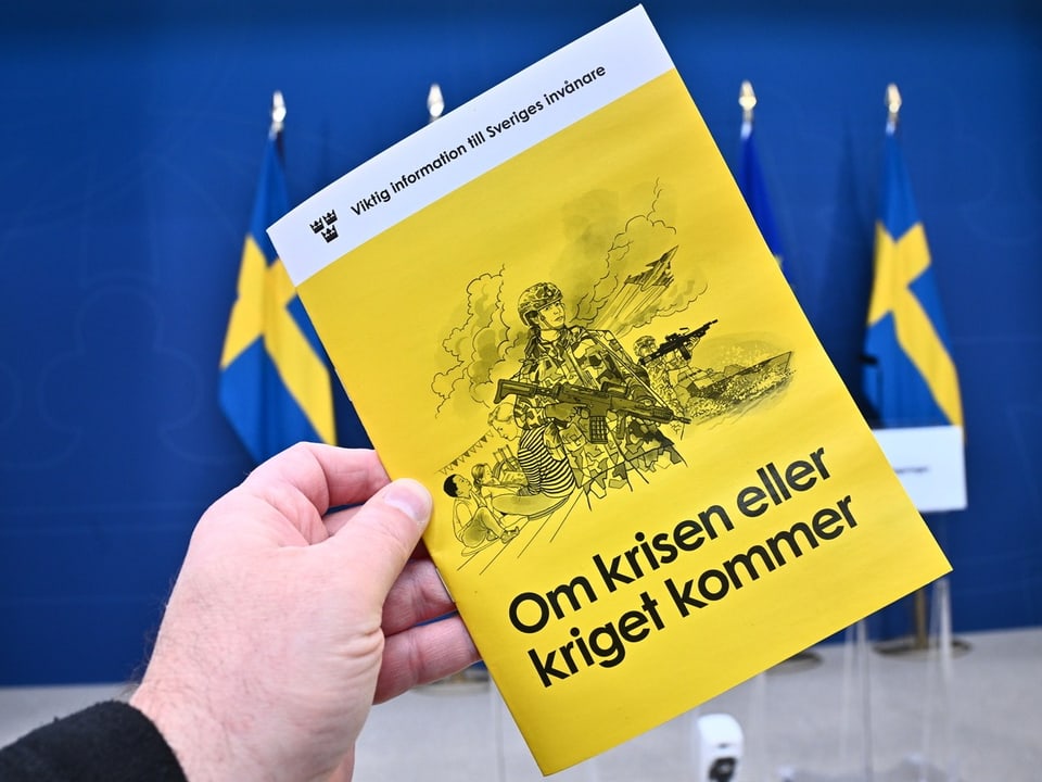 Person hält gelbes Heft mit Titel 'Om krisen eller kriget kommer' vor blauem Hintergrund mit schwedischen Flaggen.