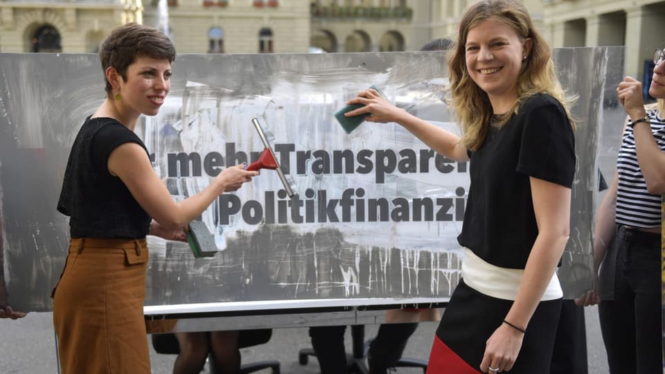 Zwei Frauen putzen ein Plakat, darauf erscheint unter dem Schmutz der Schriftzug «mehr Transparenz»
