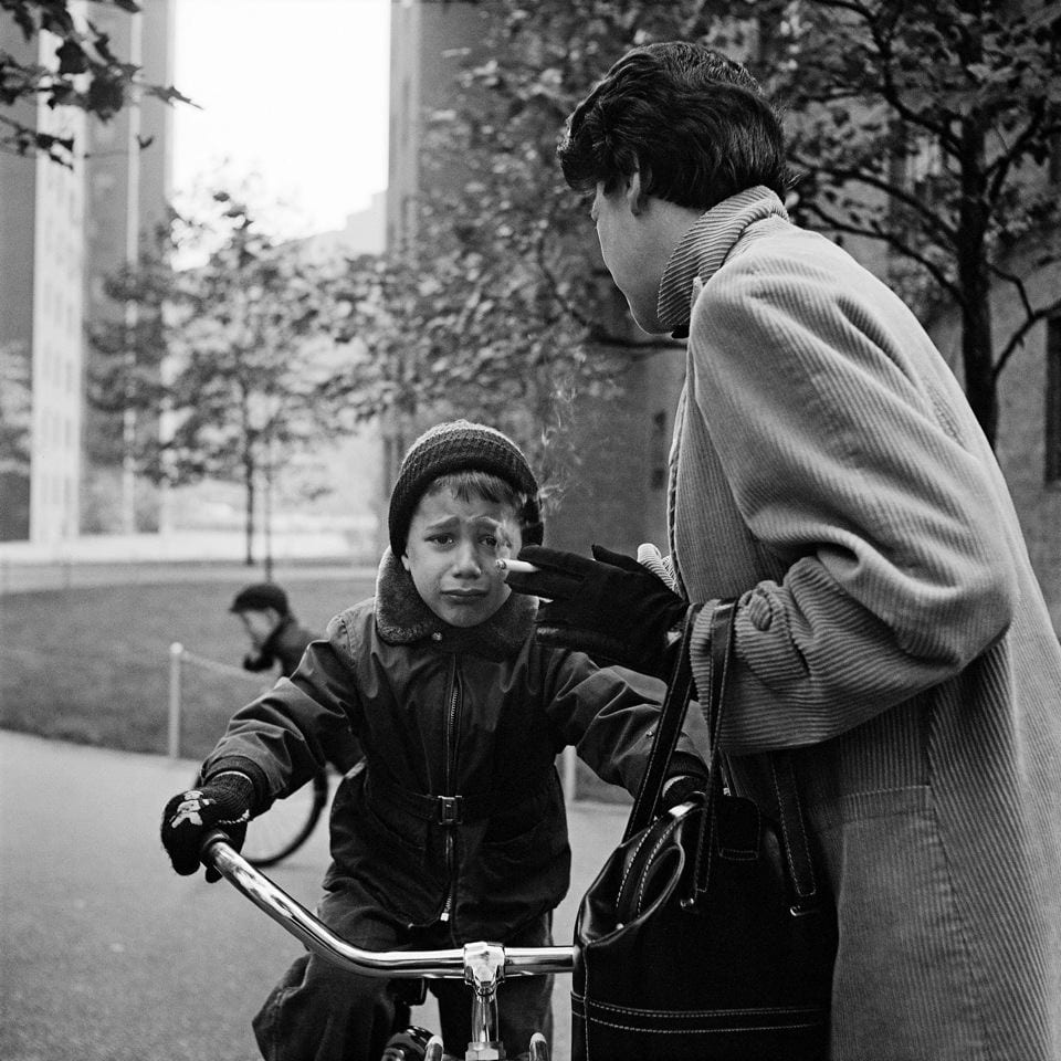 Ein weinender Junge auf einem Fahrrad. Seine Mutter steht nebenan und raucht.