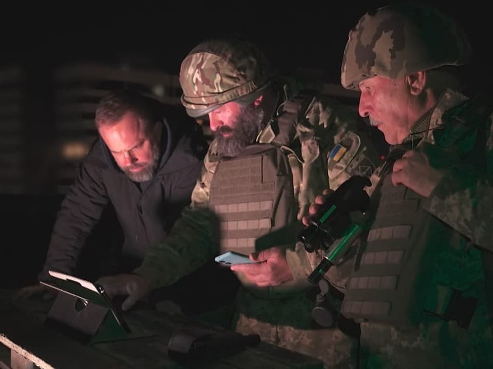 Drei Männer in militärischer Ausrüstung schauen auf ein Tablet in der Nacht.