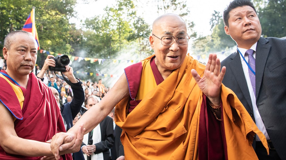 Der Dalai Lama in orangefarbenem Gewand wird begleitet von zwei Personen und winkt Menschen zu.