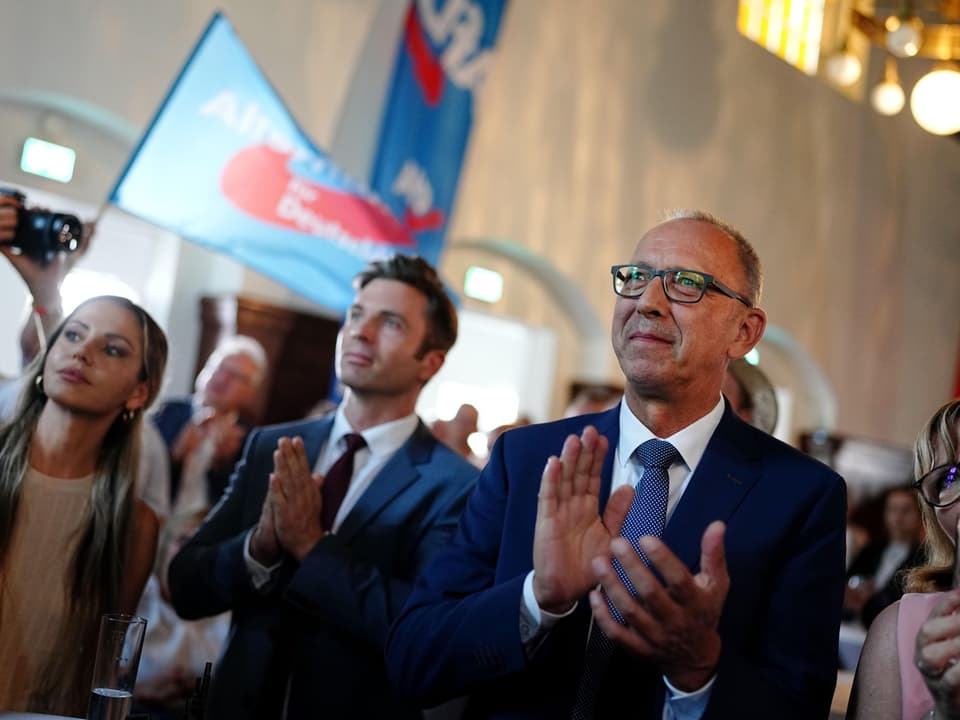 Männer klatschen in einer Halle in die Hände. AfD-Fahnen werden geschwenkt.