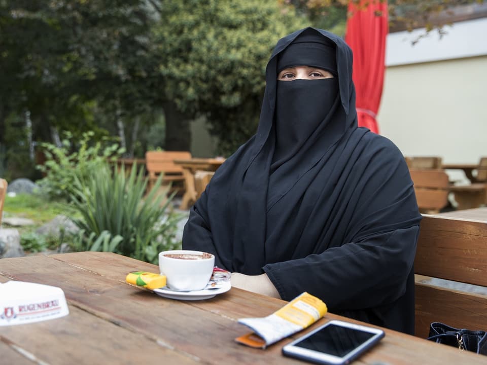 Frau sitzt mit Niqab auf einer Bank vor einem Holztisch in einem Restaurant.