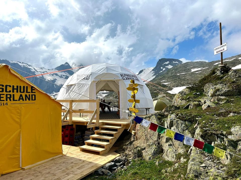 Ein gelbes und ein weisses Zelt vor einer Bergkulisse, bunte Fahnen hängen zur Dekoration.