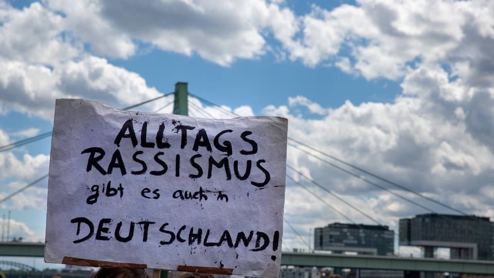 Menschenmenge von hinten, eine Person hält Schild hoch mit Schrift: Deutschland, auch du hast ein Rassismusproblem.