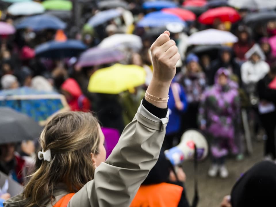 Nahaufnahme einer Person, die die Faust inmitten einer Menschenmenge mit Regenschirmen erhebt.