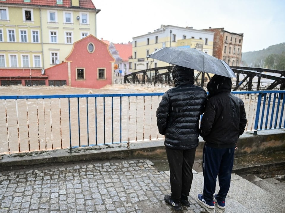 Zwei Personen unter Regenschirm, beobachten überflutete Strasse vor mehrfarbigen Gebäuden.