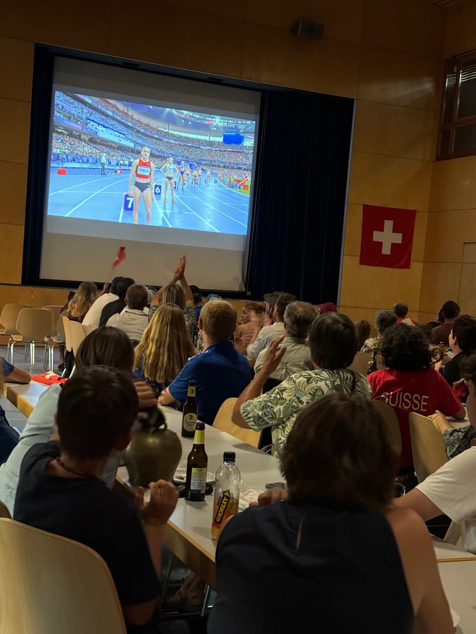 Menschen sehen ein Sportereignis auf einer Leinwand in einem Raum mit Schweizer Flagge.