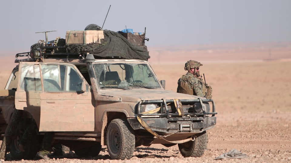 Jeep in der Wüste, daneben steht ein Mann in Kampfmontur.