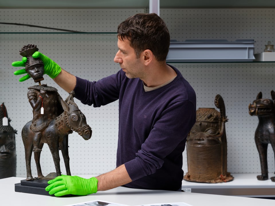 Person mit grünen Handschuhen hält afrikanische Skulptur.