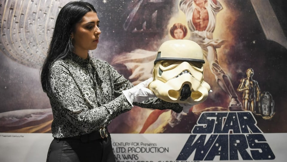 Eine Frau mit Handschuhen hält einen weissen Helm. Im Hintergrund hängt ein Star-Wars-Plakat.
