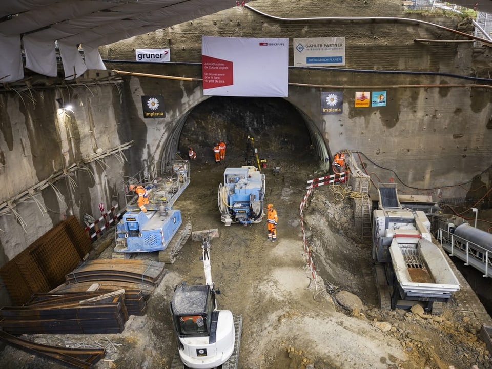 Ein paar Meter Tunnel sind in den Berg gehauen. Bauarbeiter und Baumaschinen.