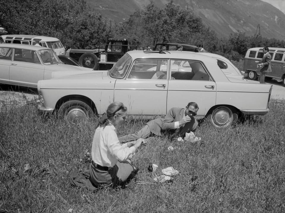 Zwei Personen picknicken auf einer Wiese neben einem Auto in den Bergen.