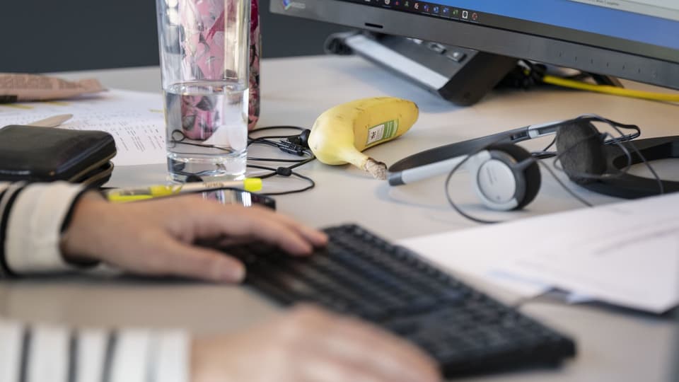 Schreibtisch mit Computer, Banane, Glas Wasser und tippender Hand.