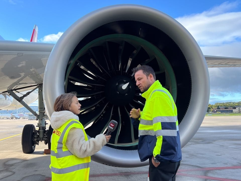 Zwei Menschen mit gelben Leuchtwesten stehen vor einem Triebwerk eines Flugzeugs. 