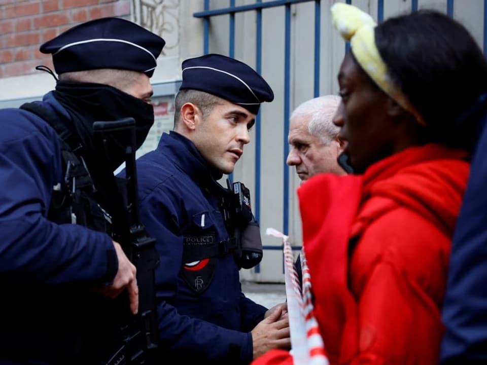 Zwei Polizisten sprechen mit Menschen hinter einer Absperrung.