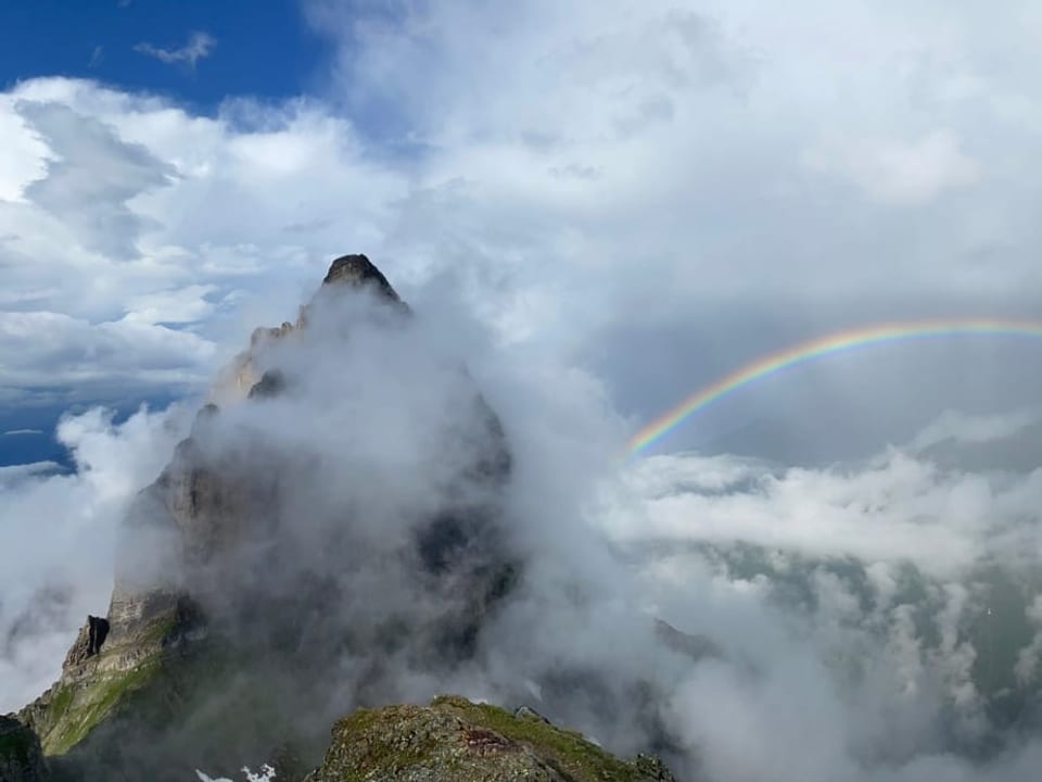 Bergwelt in Wolken, rechts ein Regenbogen, links Gipfel mit vielen Wolken.