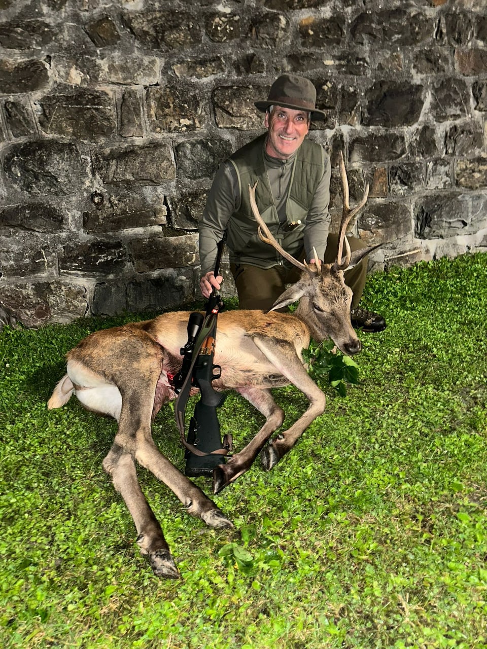 Jäger posiert mit erlegtem Hirsch vor Steinmauer.