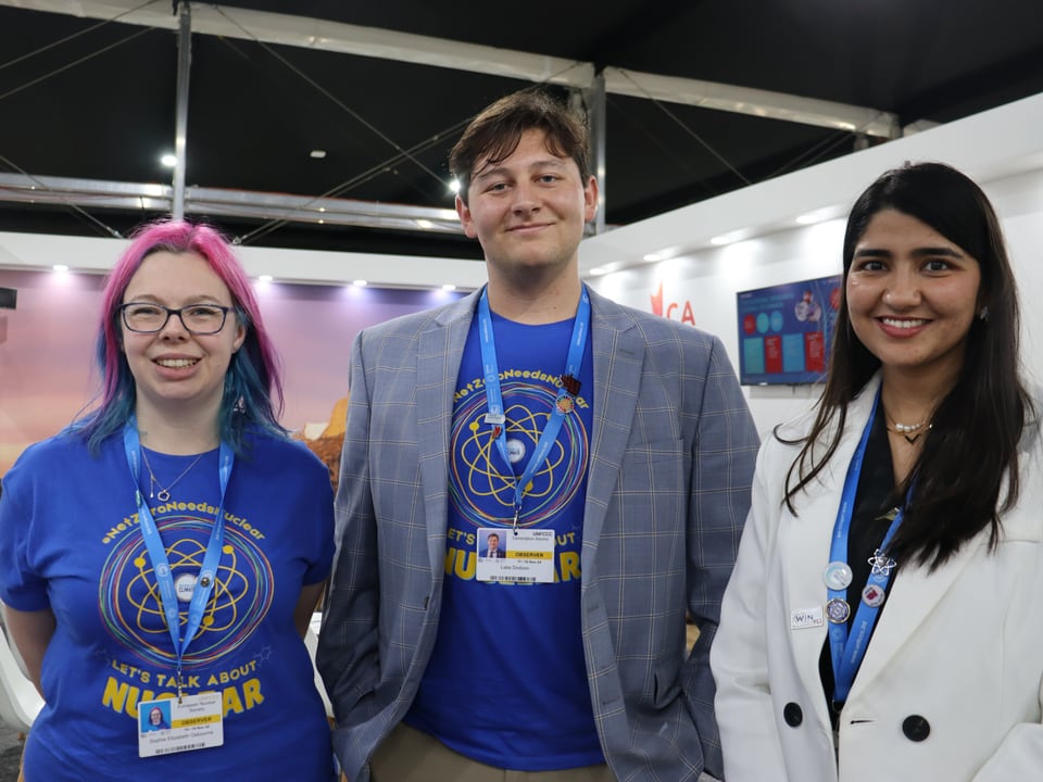 Sophie, Lake und Gunjan in blauen T-Shirs bei einer Veranstaltung in einem Messezelt.