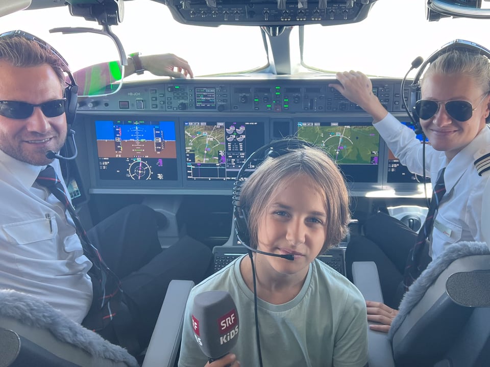 Zwei Erwachsene in Uniform und ein Junge sitzen in einem Cockpit vor vielen Bildschirmen und Knöpfen. 