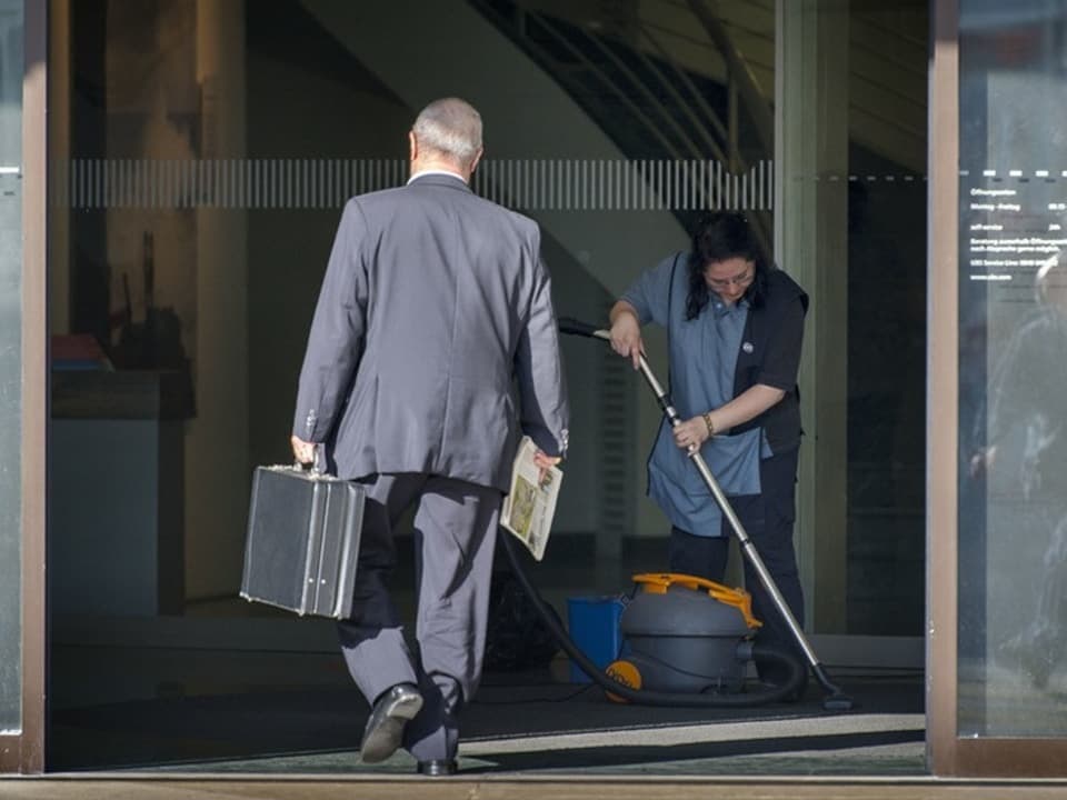 Geschäftsmann mit Koffer betritt Gebäude, Reinigungskraft beim Putzen.