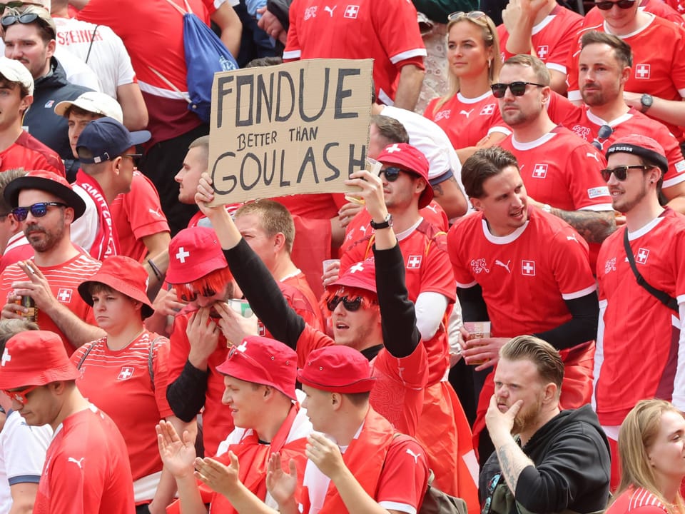 Fans in roten Trikots halten ein Schild mit der Aufschrift 'Fondue better than Goulash' hoch.