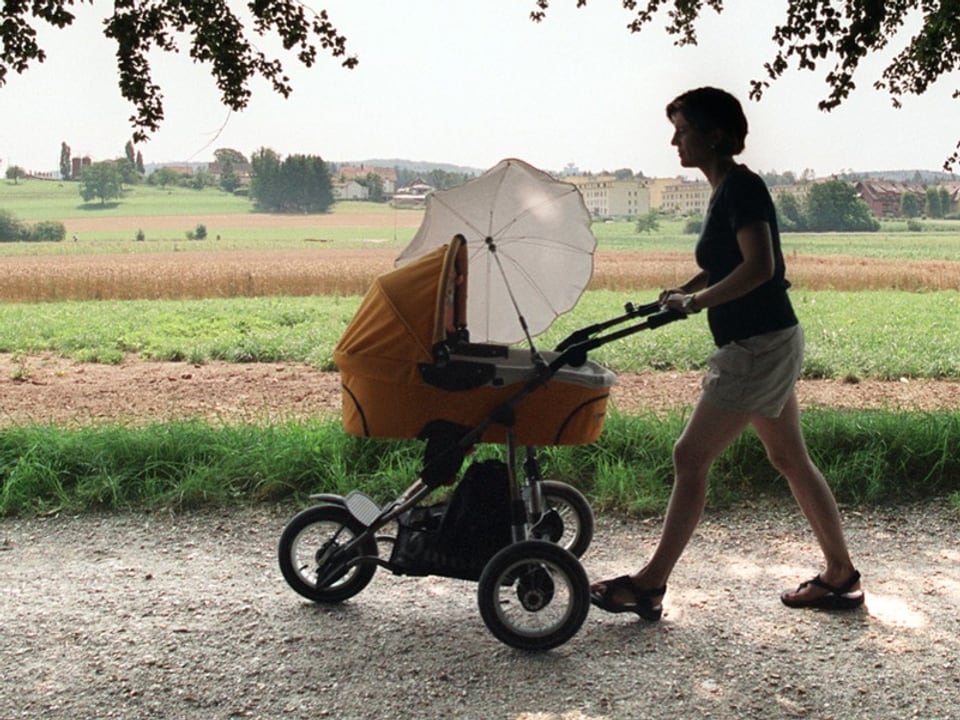 Eine Frau schiebt einen Kinderwagen auf einem Weg mit ländlicher Kulisse.