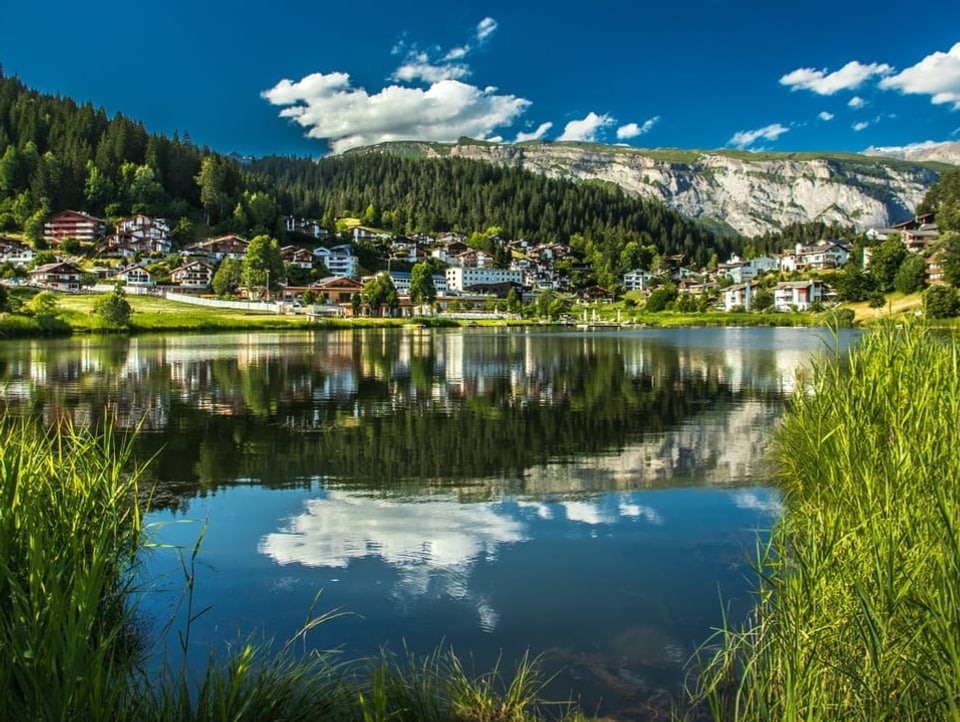Bergsee mit Dorf, in dem sich blauer Himmel spiegelt.