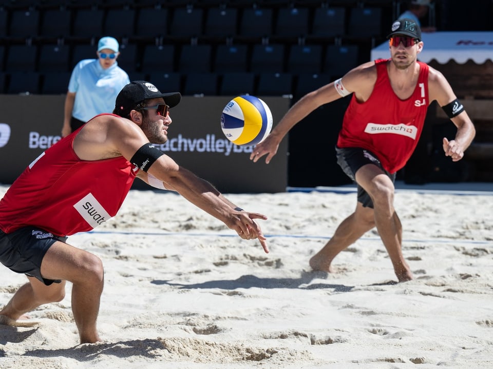 Zwei Beachvolleyballspieler in roten Trikots im Sand mit Volleyball.