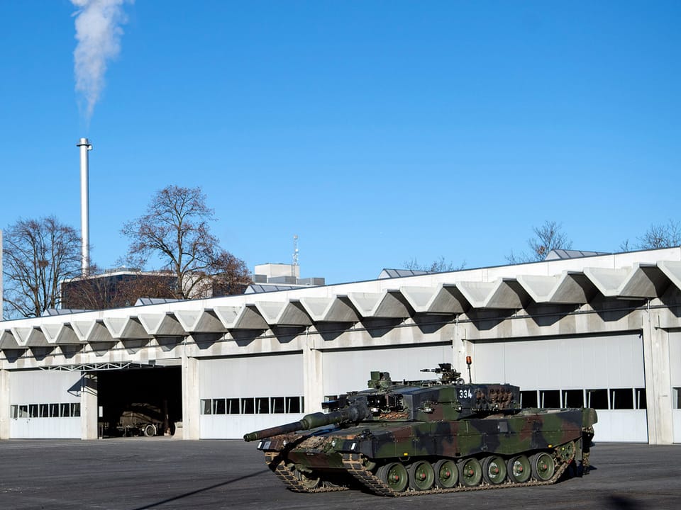 Ein Panzer steht vor einem langen, grauen Gebäude, in dem Panzer untergebracht sind.