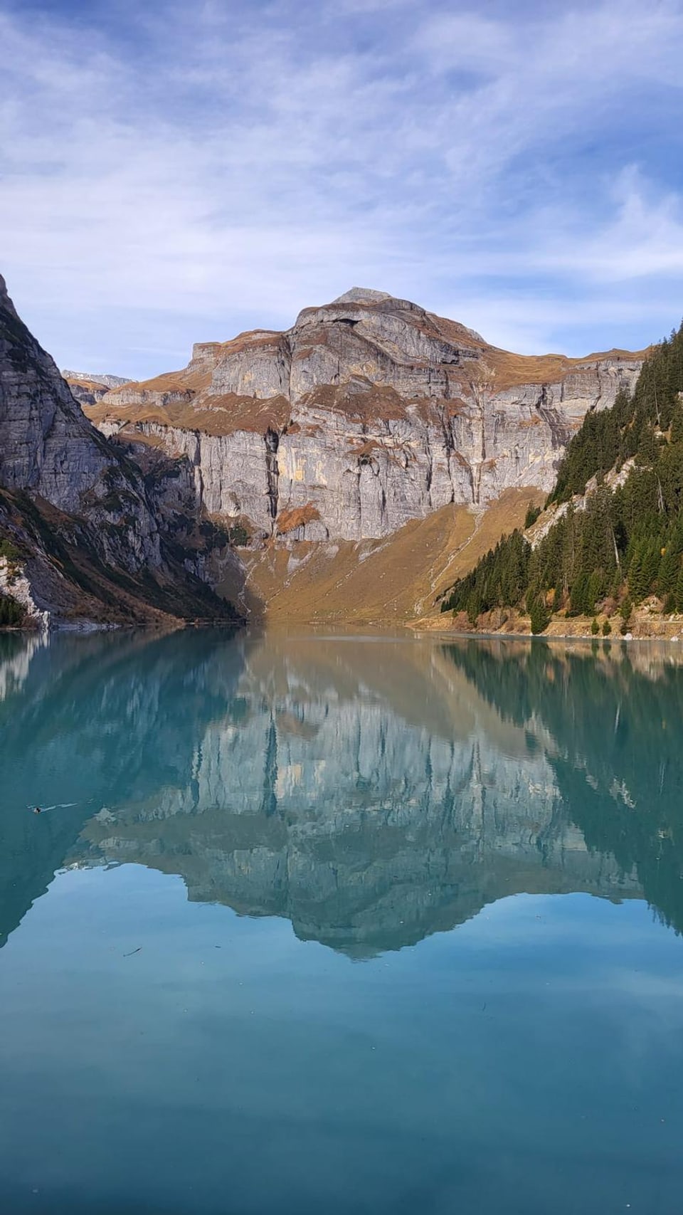 Bergsee mit Bergspiegelung und bewaldeten Hängen.