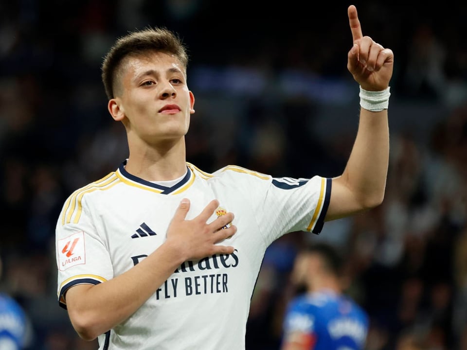 Fussballspieler in einem weissen Trikot hebt einen Finger und legt die Hand auf die Brust.