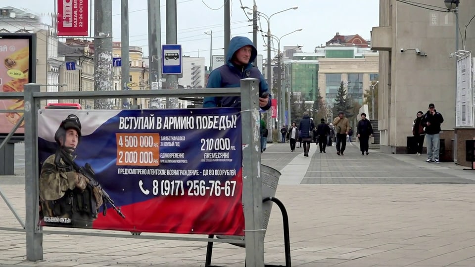 Mann steht hinter russischem Rekrutierungsplakat in Strassenansicht.