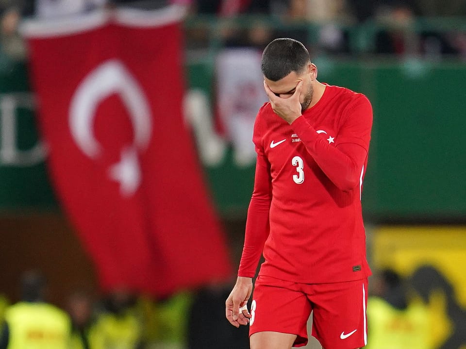 Fussballspieler in rotem Trikot bedeckt sein Gesicht, türkische Flagge im Hintergrund.