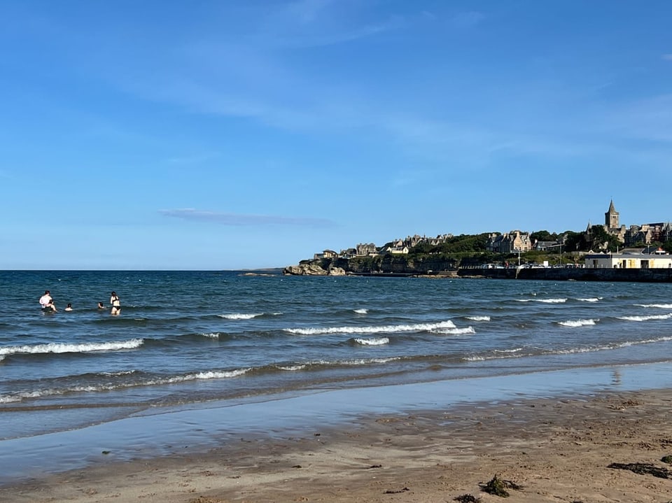 Blick auf das Meer mit zwei Personen im Wasser und einer Stadt im Hintergrund.