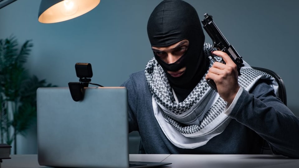 Ein Terrorist mit Maske und Pistole sitzt vor einem Laptop.