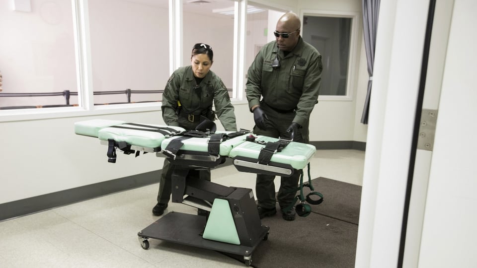 Zwei Personen in Uniform stehen bei einer Gefängnisliege in einem hellen Raum.