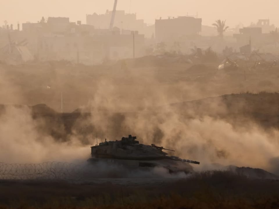 Militärpanzer bewegt sich durch staubige Landschaft vor städtischem Hintergrund.