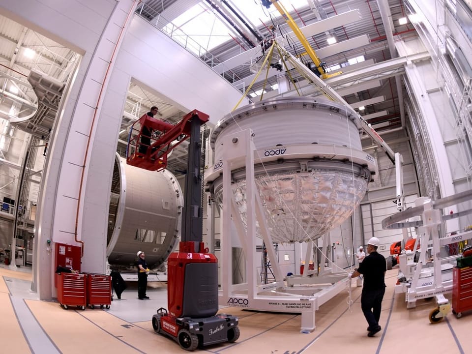 Montage einer grossen wissenschaftlichen Anlage in einer Fabrikhalle mit Kran und Arbeitern.