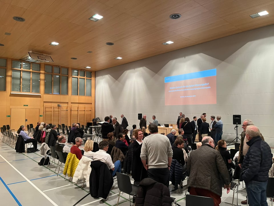 Menschen in Turnhalle bei Vortrag, leere Stühle.