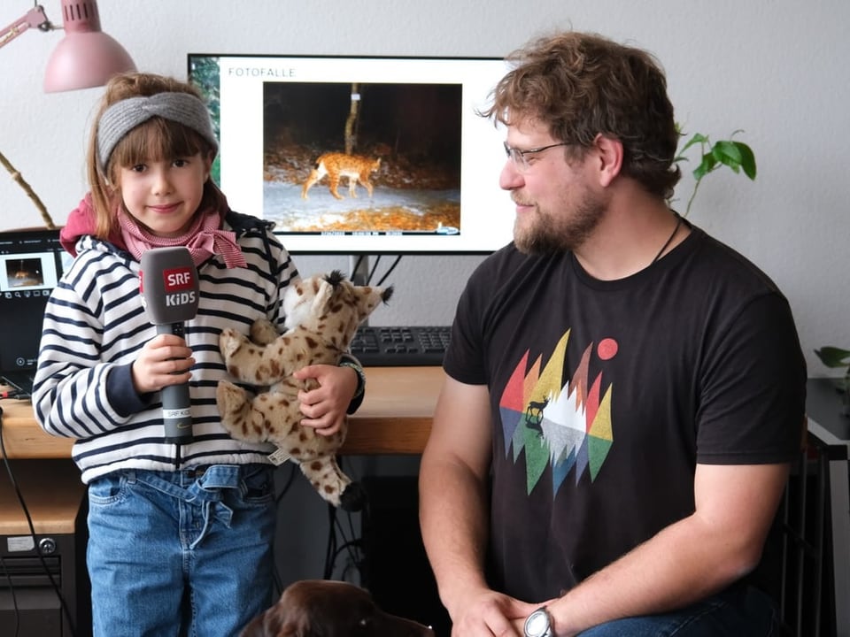 Ein Mädchen steht mit einem Mikrofon und einem Stoff-Luchs neben einem Mann. Hinter ihnen ist ein Computer. 