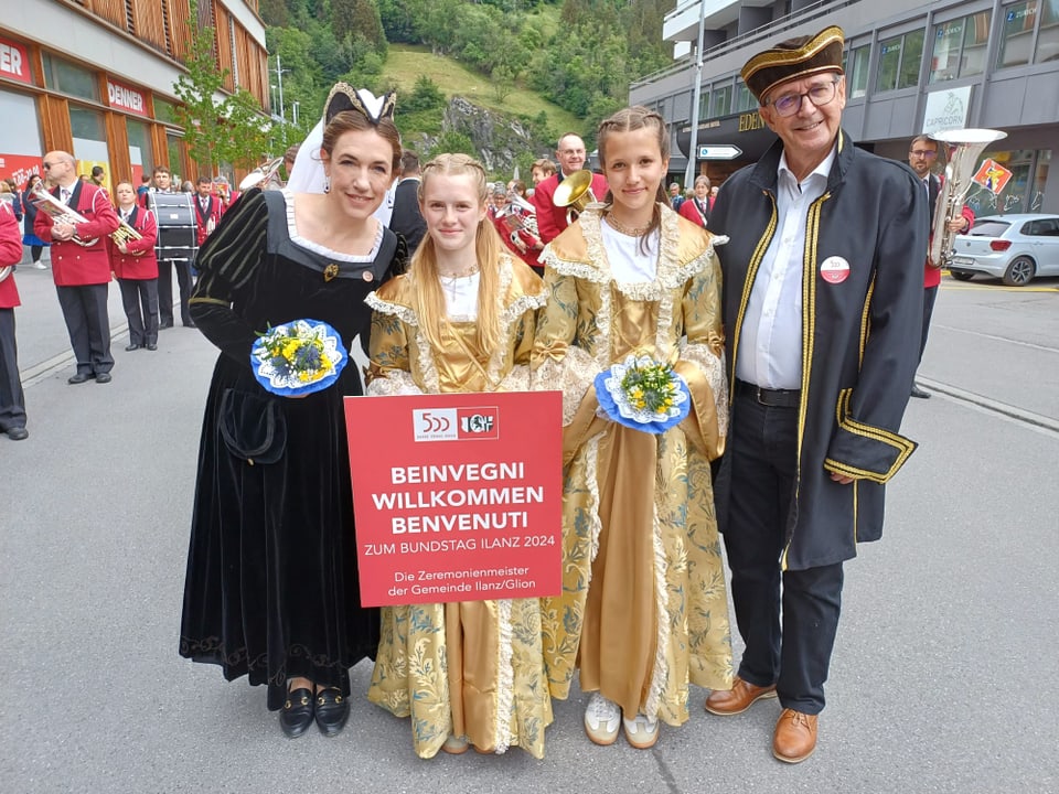 Vier Personen in historischen Gewändern halten ein Begrüssungsschild bei einer Veranstaltung im Freien.